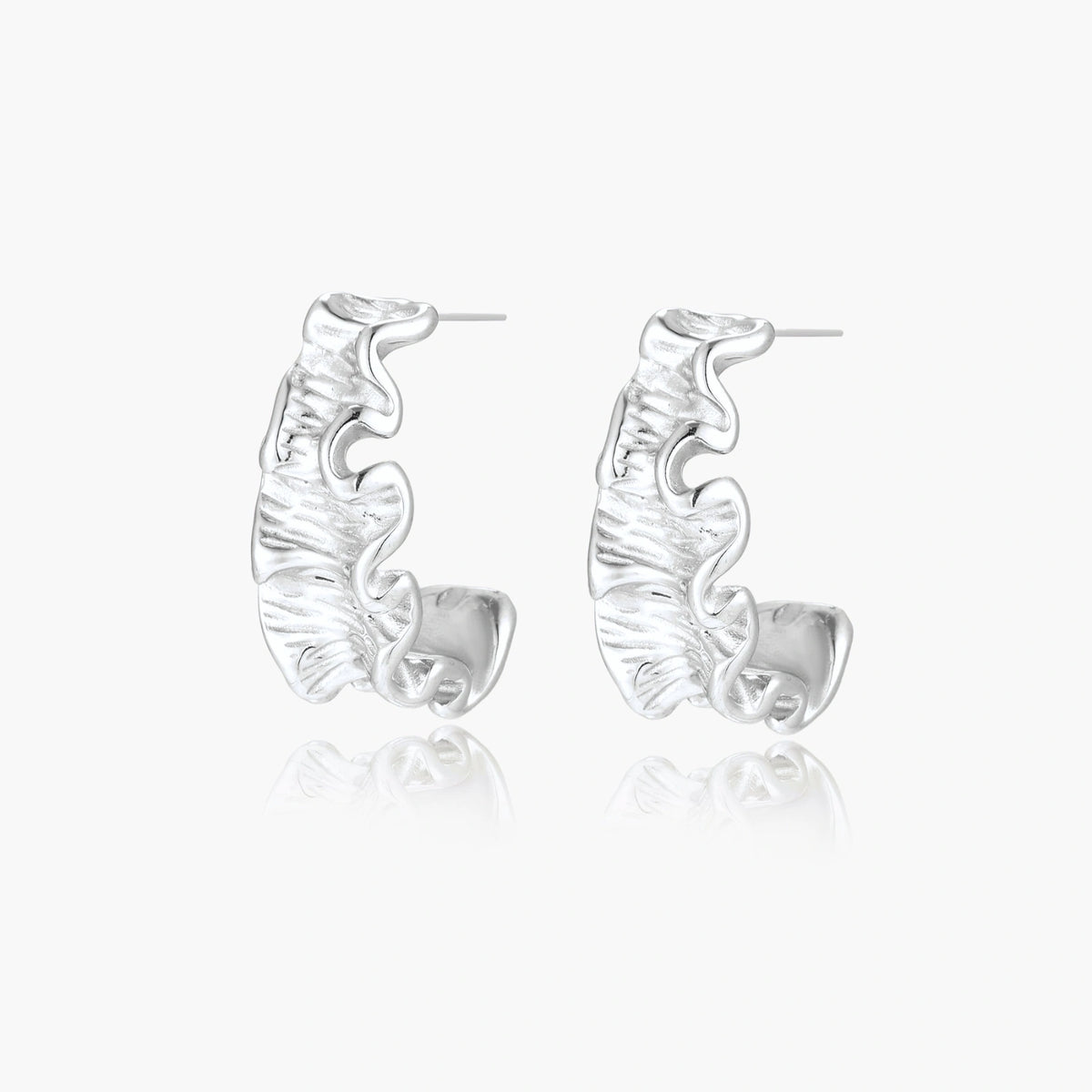 Textured white gold-plated hoop earrings with flowing wave pattern against cream background, showing dimensional rippled surface.