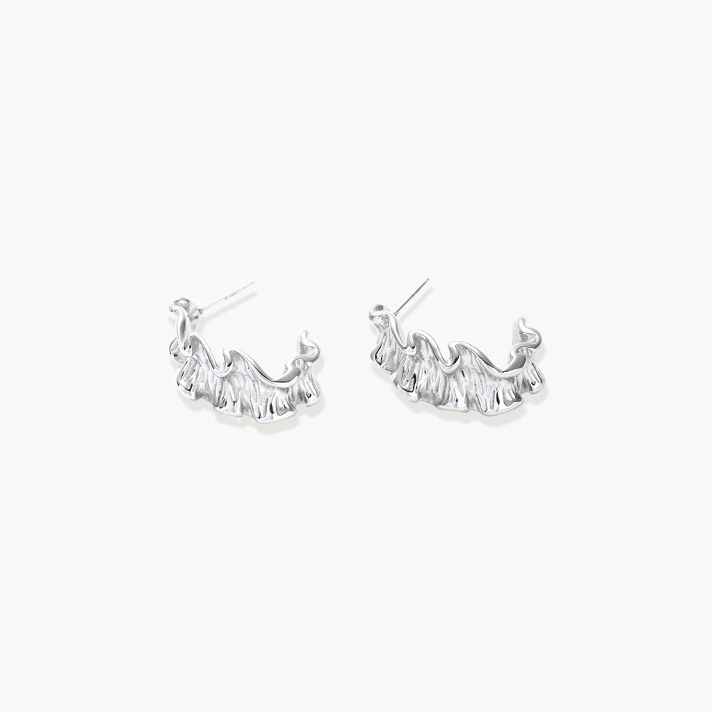 Textured white gold-plated hoop earrings with flowing wave pattern against cream background, showing dimensional rippled surface.