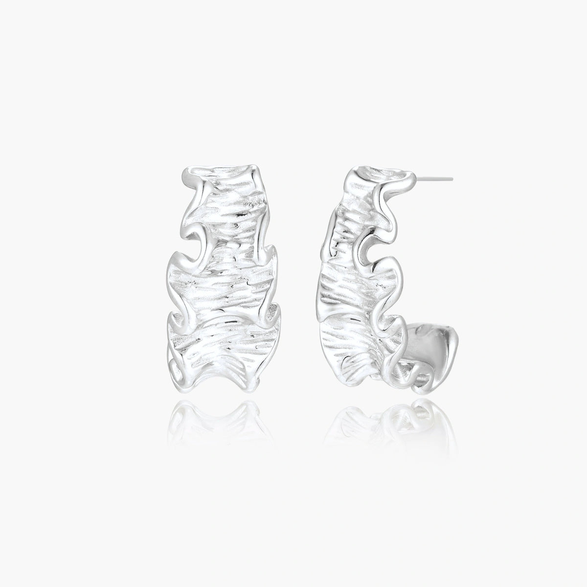 Textured white gold-plated hoop earrings with flowing wave pattern against cream background, showing dimensional rippled surface.