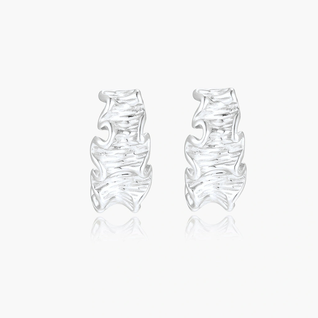 Textured white gold-plated hoop earrings with flowing wave pattern against cream background, showing dimensional rippled surface.