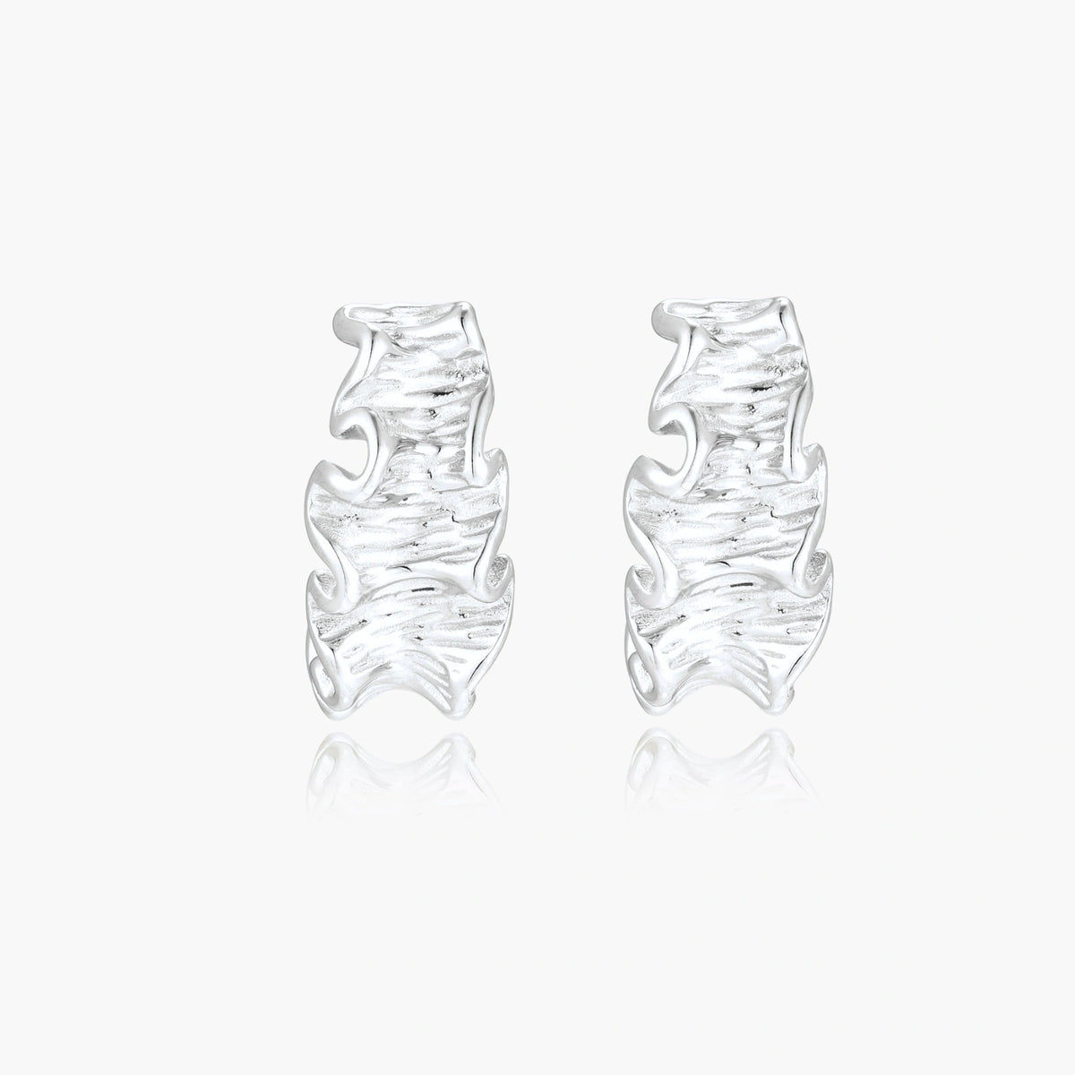 Textured white gold-plated hoop earrings with flowing wave pattern against cream background, showing dimensional rippled surface.
